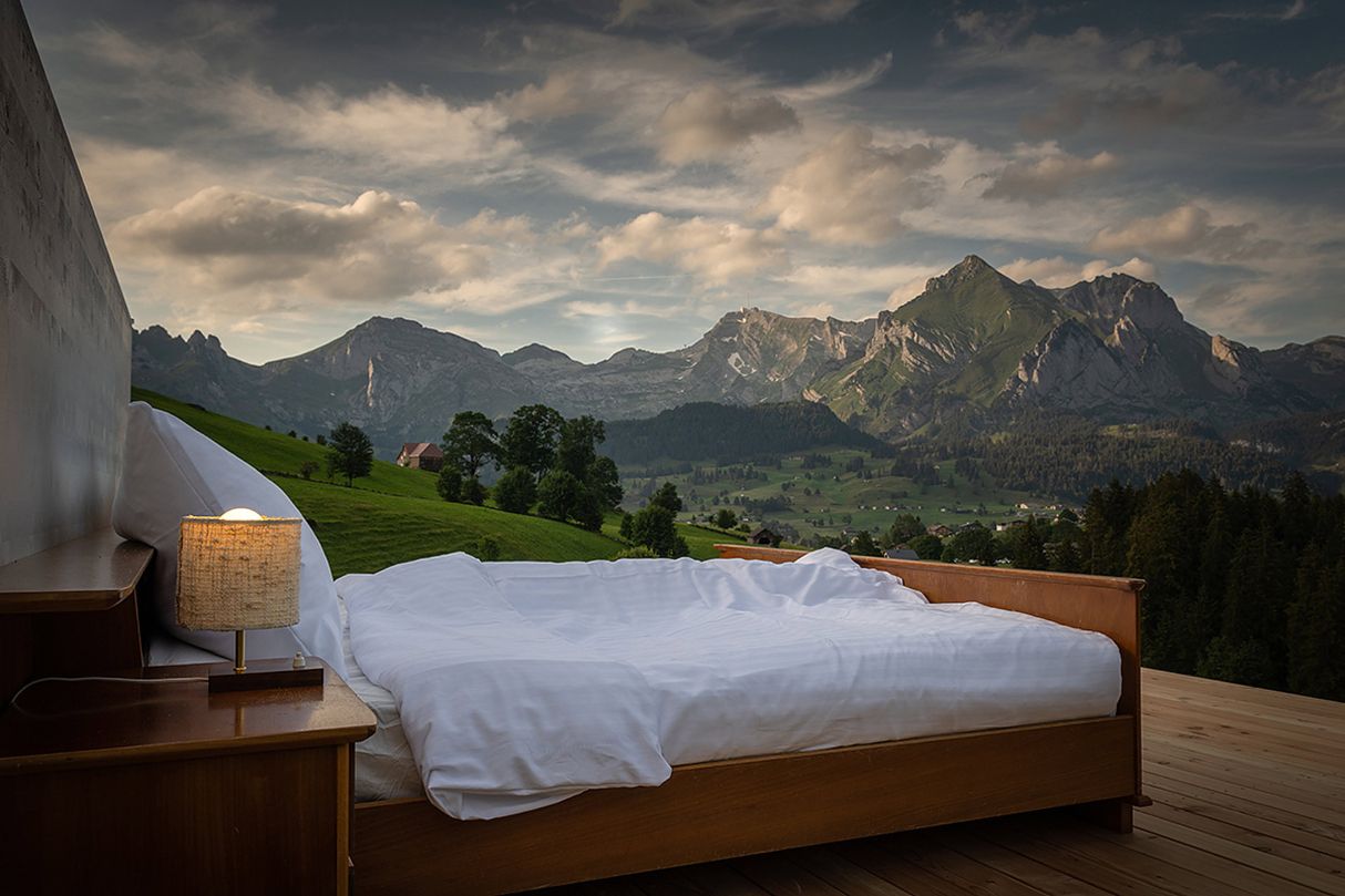 A bed in a room with windows so clean you can't see them, with a view of a mountain village at dusk